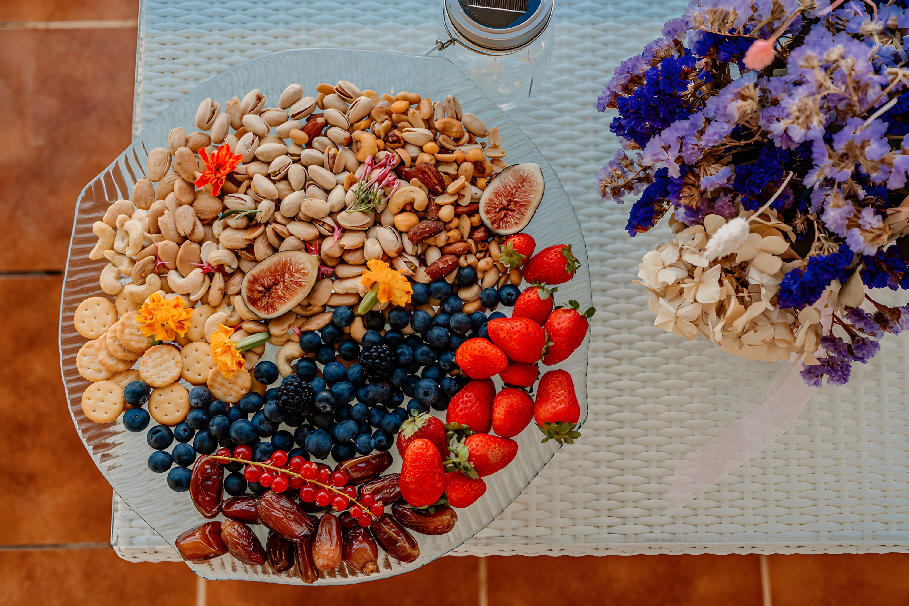 A round platter of fruit and nuts beside a bunch of purple flowers, seen from above