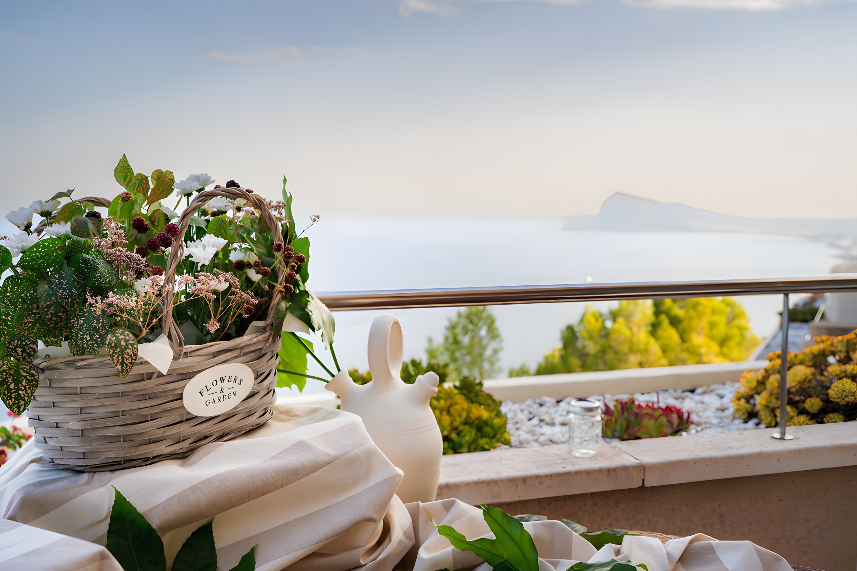 A flower basket and ceramic jug on a linen-draped terrace table above the sea